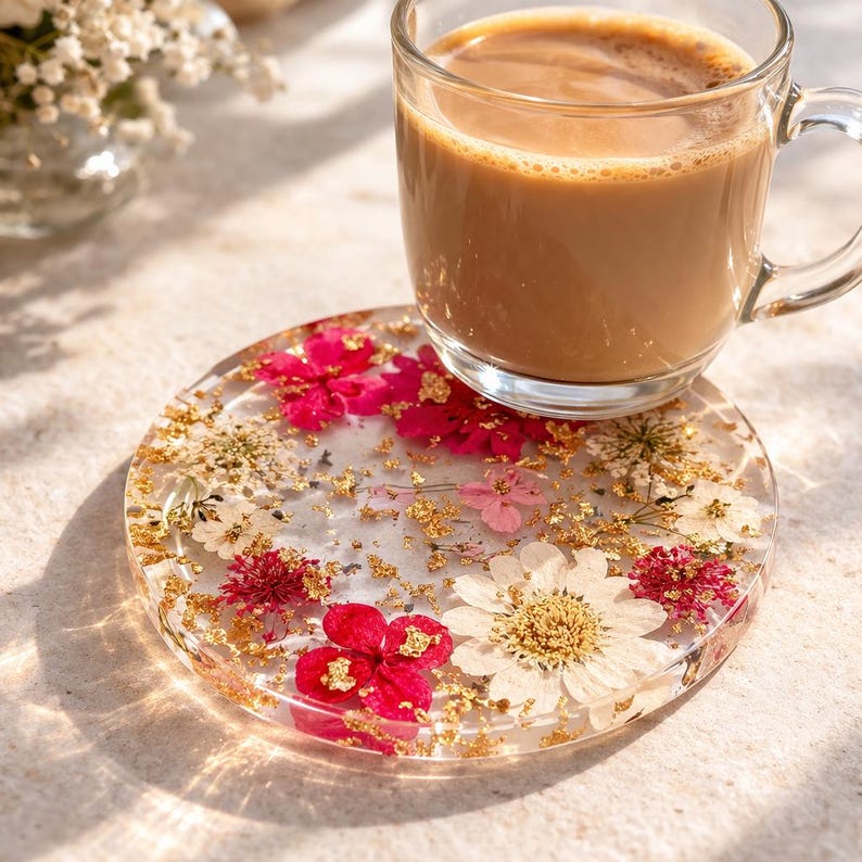 Op de afbeelding: Een heldere, ronde hars coaster met gedroogde bloemen in rood, roze en wit, met goudblad accenten. Een glazen mok met een lichtbruine drank staat op de coaster. De coaster heeft een diameter van ongeveer 10 cm.