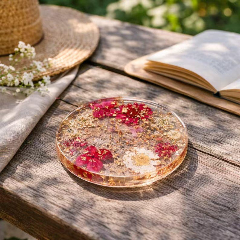 Op de afbeelding: Een heldere, ronde hars coaster met ingebedde rode en witte bloemen en gouden vlokken. De coaster ligt op een houten oppervlak, met een strohoed, boek en bloemen op de achtergrond.