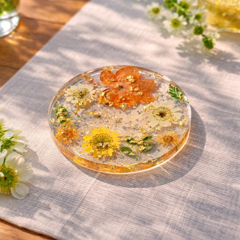 Op de afbeelding: Een heldere, ronde hars coaster met ingebedde gedroogde bloemen en bladgoudvlokken. De coaster heeft gele, oranje en witte bloemen, wat een decoratief ontwerp cre&euml;ert. Perfect voor interieurdecoratie.