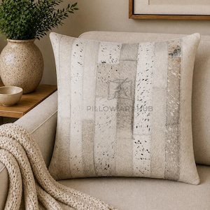 May include: A square throw pillow featuring a patchwork design of light beige, cream, and grey cowhide strips with natural speckled patterns. The center of the pillow displays a faint watermark logo with the text PILLOWAR HUB in capital letters.