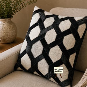 Può includere: Un cuscino quadrato decorativo con un motivo geometrico di forme nere e bianche. Il cuscino è realizzato in un materiale strutturato ed è posto su una poltrona beige. Il logo The Pillow Art Hub è visibile.