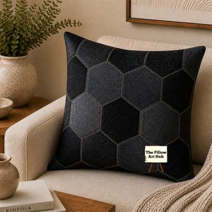 Può includere: Un cuscino nero decorativo con un motivo geometrico di esagoni grigio scuro e neri. Il cuscino è su un divano beige con una coperta beige. Un tavolino in legno contiene un vaso con vegetazione e una piccola ciotola.