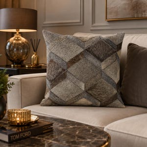 May include: A square throw pillow featuring a geometric patchwork design made of textured animal hide in shades of gray, brown, and cream. The pattern consists of repeating diamond and triangular shapes, creating a multi-dimensional visual effect.