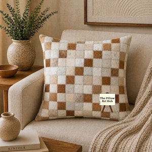 Puede incluir: Cojín decorativo con diseño de patchwork cuadrado en tonos marrón, crema y beige. El cojín está sobre un sillón beige. Una manta de punto beige está sobre el brazo de la silla.