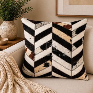 May include: A square throw pillow featuring a chevron pattern made of stitched panels of cowhide. The design consists of alternating strips of solid black, white, and brown-spotted fur, creating a textured, geometric appearance on the surface.