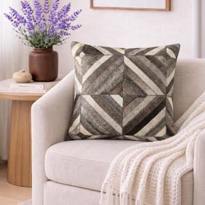 May include: A square cushion with a geometric design in grey, brown, and cream tones. It rests on a cream armchair, with a cream throw blanket. A vase of purple flowers is in the background. The cushion's pattern is made of various shapes.