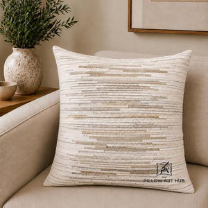 May include: A square throw pillow featuring a horizontal striped pattern in shades of cream, beige, and light brown. The surface has a textured, layered appearance. The bottom right corner displays a logo with the text THE PILLOW ART HUB.