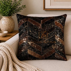 Può includere: Un cuscino quadrato con un motivo a spina di pesce composto da strisce alternate di pelle animale nera e marrone con macchie dorate metallizzate. Al centro del cuscino è presente un logo tenue con il testo PILLOW ART HUB.
