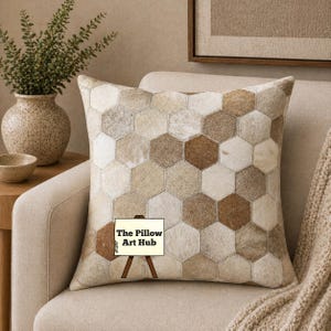 May include: A square decorative pillow with a geometric pattern of hexagonal shapes in shades of beige, brown, and cream. The pillow is made of a textured material and rests on a beige armchair. The Pillow Art Hub is written on a sign.