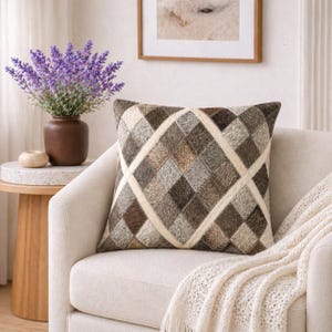 Peut inclure: Coussin carré décoratif avec un motif géométrique dans les tons de marron, gris et crème. Le coussin est posé sur un fauteuil crème, avec une couverture crème drapée sur le bras. Un vase de fleurs violettes est sur une table d'appoint.