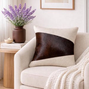 May include: A square decorative pillow with a cream-colored fabric and a diagonal panel of dark brown cowhide. The pillow is placed on a cream-colored armchair, with a white blanket draped over the arm.
