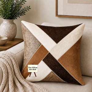 May include: Square decorative pillow with a geometric patchwork design in shades of brown, beige, and cream. The pillow is on a beige sofa, with a beige throw blanket and a vase of greenery in the background. The Pillow Art Hub logo is visible.
