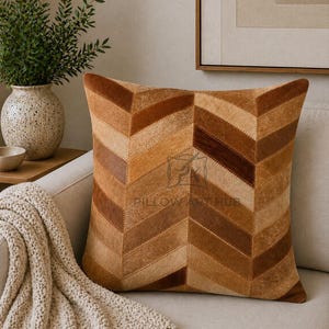 May include: A square throw pillow featuring a chevron pattern made of stitched panels of hair-on hide in various shades of brown, tan, and cream. The center of the pillow displays a faint watermark logo reading PILLOW ART HUB.