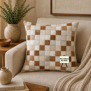 Peut inclure: Coussin décoratif à motifs carrés dans les tons marron, crème et beige. Le coussin est posé sur un fauteuil beige. Un plaid en tricot beige est drapé sur le bras du fauteuil.