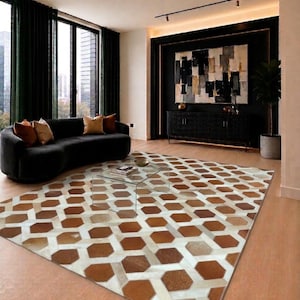 Op de afbeelding: Een rechthoekig vloerkleed met een geometrisch patroon van in elkaar grijpende zeshoeken in bruin-, beige- en wittinten. Het kleed is opgebouwd uit individuele stukken huid met een textuur, wat een herhalend mozaïekontwerp creëert.
