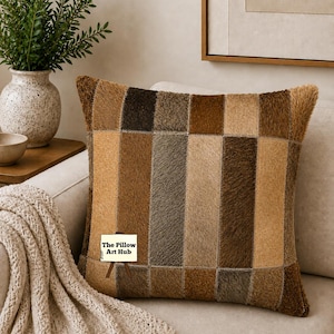 Può includere: Un cuscino decorativo quadrato con un design patchwork nei toni del marrone, beige e grigio. Il cuscino è su un divano beige, con una coperta color crema e un vaso con piante verdi sullo sfondo. Il logo The Pillow Art Hub è visibile.