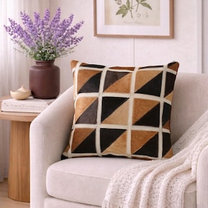 Pode incluir: Almofada decorativa quadrada com um padrão geométrico de triângulos castanhos, pretos e bege. A almofada está em uma poltrona creme, com um vaso de flores roxas e uma impressão botânica emoldurada no fundo.