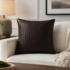 May include: A square throw pillow featuring a dark brown leather exterior with a woven, basket-weave pattern across the front panel. The pillow has a smooth, solid leather border framing the intricate, interlaced texture of the central design.