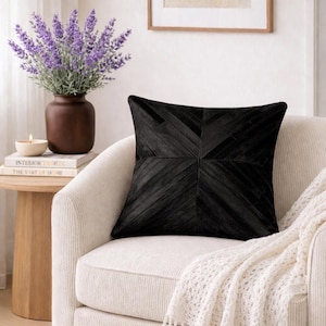 May include: A black decorative pillow with a geometric design rests on a cream-colored armchair. The pillow has a textured surface and is made of a soft material. A white knitted throw blanket is draped over the chair.