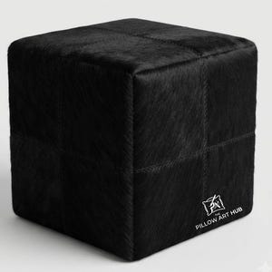 Puede incluir: Un taburete de forma cúbica en color negro tapizado en cuero con pelo texturizado. La superficie presenta una cuadrícula de costuras visibles. Un logotipo blanco con el texto The Pillow Art Hub está impreso en la parte inferior derecha.