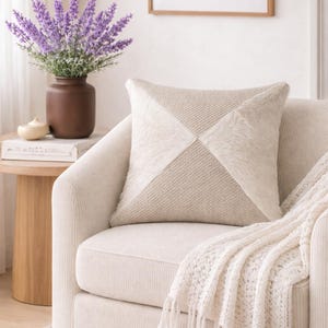 Op de afbeelding: Een crèmekleurig vierkant kussen met een geometrisch ontwerp ligt op een bijpassende fauteuil. Het kussen heeft delen van textiel en imitatiebont. Een crèmekleurige gebreide deken met franjes is over de arm gedrapeerd. Een vaas met lavendel staat in de buurt.