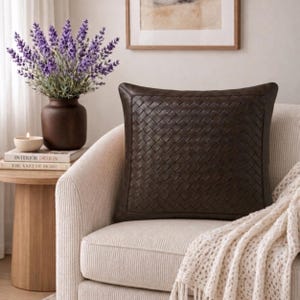 May include: A square throw pillow featuring a dark brown woven leather-like texture on the front surface. The pillow has a solid border frame and is positioned on a light-colored upholstered chair with a cream-colored knit throw blanket draped over it.