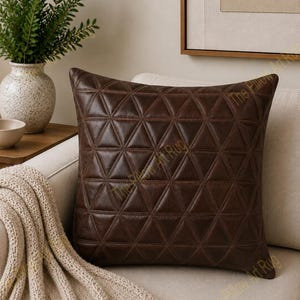 May include: A square dark brown pillow featuring a geometric pattern of stitched triangles across the entire surface. The pillow has a smooth texture and is positioned upright, showcasing a consistent grid of equilateral triangles created by seams.