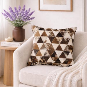 Può includere: Cuscino decorativo quadrato con un motivo geometrico di triangoli marroni, beige e bianchi. Il cuscino è su una poltrona color crema. Un vaso di fiori viola e un tavolino sono sullo sfondo.