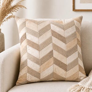 May include: Square decorative pillow with a chevron pattern in shades of beige and brown. The pillow is made of a textured material and is placed on a cream-colored sofa. The pillow is a home decor item.