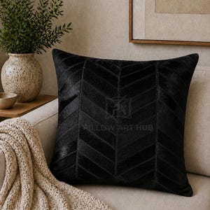 May include: A square black throw pillow featuring a chevron pattern made of stitched panels of hair-on hide. The center of the pillow displays a faint watermark logo with the text PILLOW ART HUB. The texture appears smooth and directional.