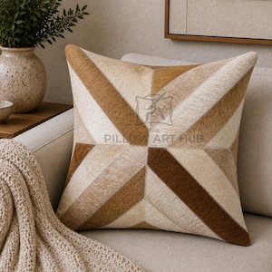 May include: A square throw pillow featuring a geometric pattern of alternating light cream, tan, and dark brown hair-on-hide panels. The center of the pillow displays a faint watermark logo with the text THE PILLOW ART HUB.