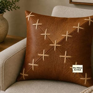 May include: A square, brown leather pillow with a stitched cross pattern. The pillow is on a beige armchair. The Pillow Art Hub logo is visible in the lower right corner. A vase with greenery and a wooden bowl are in the background.