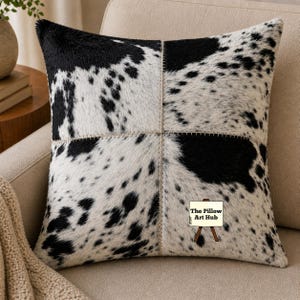 May include: Square decorative pillow with a cowhide pattern in black and white. The pillow is divided into four sections, stitched together with a light-colored thread. The Pillow Art Hub logo is visible on the pillow.