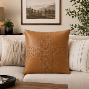 May include: A square throw pillow in a tan brown color featuring a central cross design composed of woven leather strips. The pillow has smooth leather panels on the sides and top and bottom, creating a textured and structured appearance.