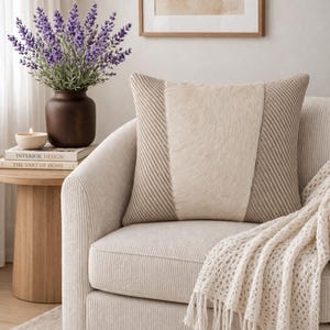 May include: A decorative pillow with a central panel of light-colored fur flanked by textured, patterned fabric. The pillow rests on a cream-colored armchair, complemented by a fringed throw blanket. A vase of lavender and a small side table complete the cozy scene.