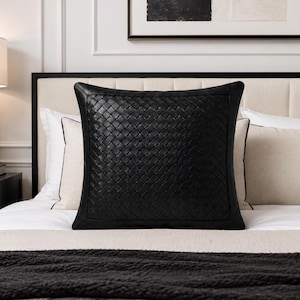 May include: A square throw pillow featuring a black woven leather-like texture on the front panel with a smooth black border. The pillow is centered and displayed in a bedroom setting, showcasing a detailed grid-like pattern across its surface.