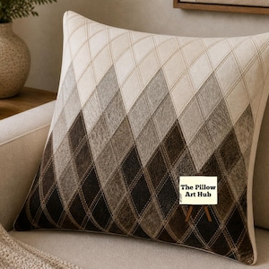Op de afbeelding: Een decoratief vierkant kussen met een geometrisch ruitpatroon in beige, bruin en zwart. Het kussen is gemaakt van een textuurrijk materiaal en ligt op een beige bank. Het logo van The Pillow Art Hub is zichtbaar.