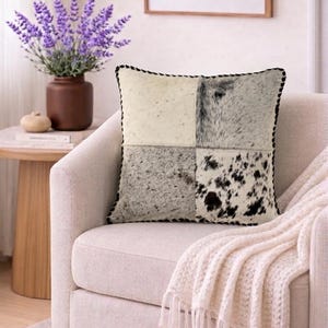 May include: Square decorative pillow with a patchwork design. The pillow features four sections of cowhide in shades of cream, gray, and black, with a black stitched border. It rests on a cream-colored armchair, with a cream-colored throw blanket.