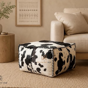 May include: A square floor pouf featuring a black and white cowhide pattern with thick braided black and white piping along the edges. The text THE PILLOW ART HUB is printed in small letters on the side of the pouf and in the bottom left corner.