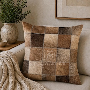 May include: A square throw pillow featuring a patchwork design of brown, tan, and cream-colored hair-on hide squares. The pillow has visible stitching between each square and includes the text PILLOW ART HUB printed in white on the front surface.