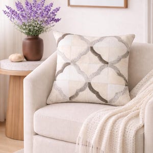 Può includere: Un cuscino decorativo con un motivo geometrico nei toni del crema, grigio e marrone poggia su una poltrona color crema. Una coperta color crema con frange è drappeggiata sul braccio. Un vaso di fiori viola è su un tavolino.
