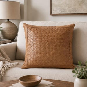 May include: A square throw pillow featuring a tan, woven leather-like front panel with a basketweave pattern, framed by a smooth, solid tan border. The pillow has a structured, flat design and is centered on a light-colored upholstered sofa.