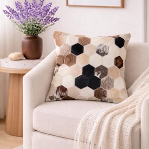 May include: A decorative throw pillow with a geometric pattern of hexagonal shapes in various shades of brown, beige, and black. The pillow is placed on a cream-colored armchair, with a cream-colored throw blanket draped over the arm.