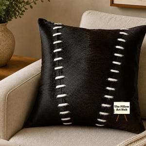May include: A square black decorative pillow with white stitching details. The pillow is made of a textured material and is placed on a beige armchair. The Pillow Art Hub logo is visible on the pillow.