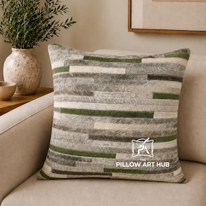 Può includere: Un cuscino quadrato con un design patchwork a strisce orizzontali nei toni del grigio, bianco e verde bosco. La superficie ha un aspetto testurizzato simile al pelo. Il testo THE PILLOW ART HUB è stampato in bianco in basso a destra.