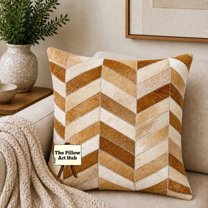 May include: Square decorative pillow with a chevron pattern in shades of brown and cream. The pillow is made of a textured material and has a stitched design. The Pillow Art Hub is written on a small sign in front of the pillow.