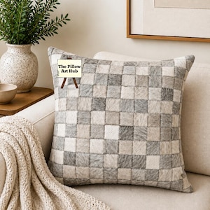 Puede incluir: Cojín decorativo cuadrado con un diseño de patchwork en tonos grises y blancos. El cojín está sobre un sofá beige con una manta color crema. El logotipo de The Pillow Art Hub es visible.