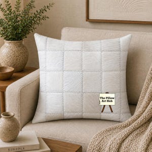 Può includere: Un cuscino decorativo quadrato bianco con un design patchwork, appoggiato su una poltrona beige. Il cuscino ha un aspetto strutturato, simile alla pelliccia, ed è cucito con cuciture visibili. Il logo The Pillow Art Hub è visibile.