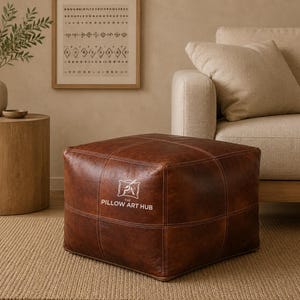 May include: A square, brown leather ottoman featuring visible stitched seams and a white logo printed on the front that reads THE PILLOW ART HUB with a small graphic of a pillow above the text. The ottoman has a smooth, textured surface.