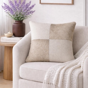 May include: A square cushion with a patchwork design in cream and beige. The cushion is on a cream armchair, with a white fringed throw draped over the arm. A vase of purple flowers sits on a side table next to the armchair.
